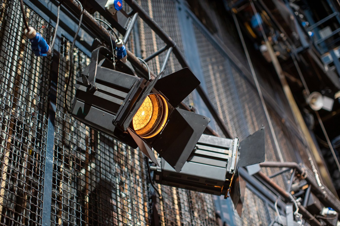 A theatrical lighting fixture with a honeycomb lens glowing warm amber against wire mesh catwalk.