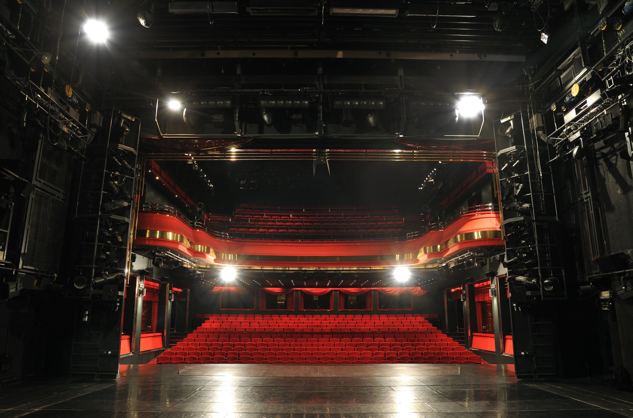 A view from the stage of an empty theater, tiered red seating rising toward the back, gold trim on the balcony and dark lighting rigging overhead.