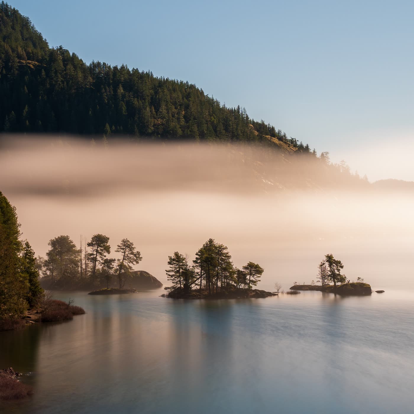 A misty mountain coastline at dawn with small pine-covered islands reflected in still water.